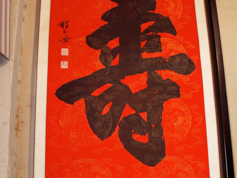 Une de ses calligraphies en très grand format : le caractère « longévité », exécuté sur un papier rouge. Ce caractère est fréquemment traité en calligraphie. Un ouvrage chinois en collige plus de 300 versions de différents calligraphes, styles et époques. C’est un document efficace pour montrer l’extrême diversité des interprétations d’un même caractère.